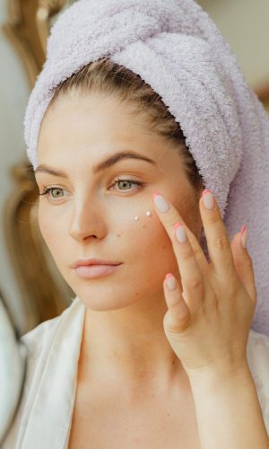 Woman with head towel applying moisturizer in front of mirror indoors.